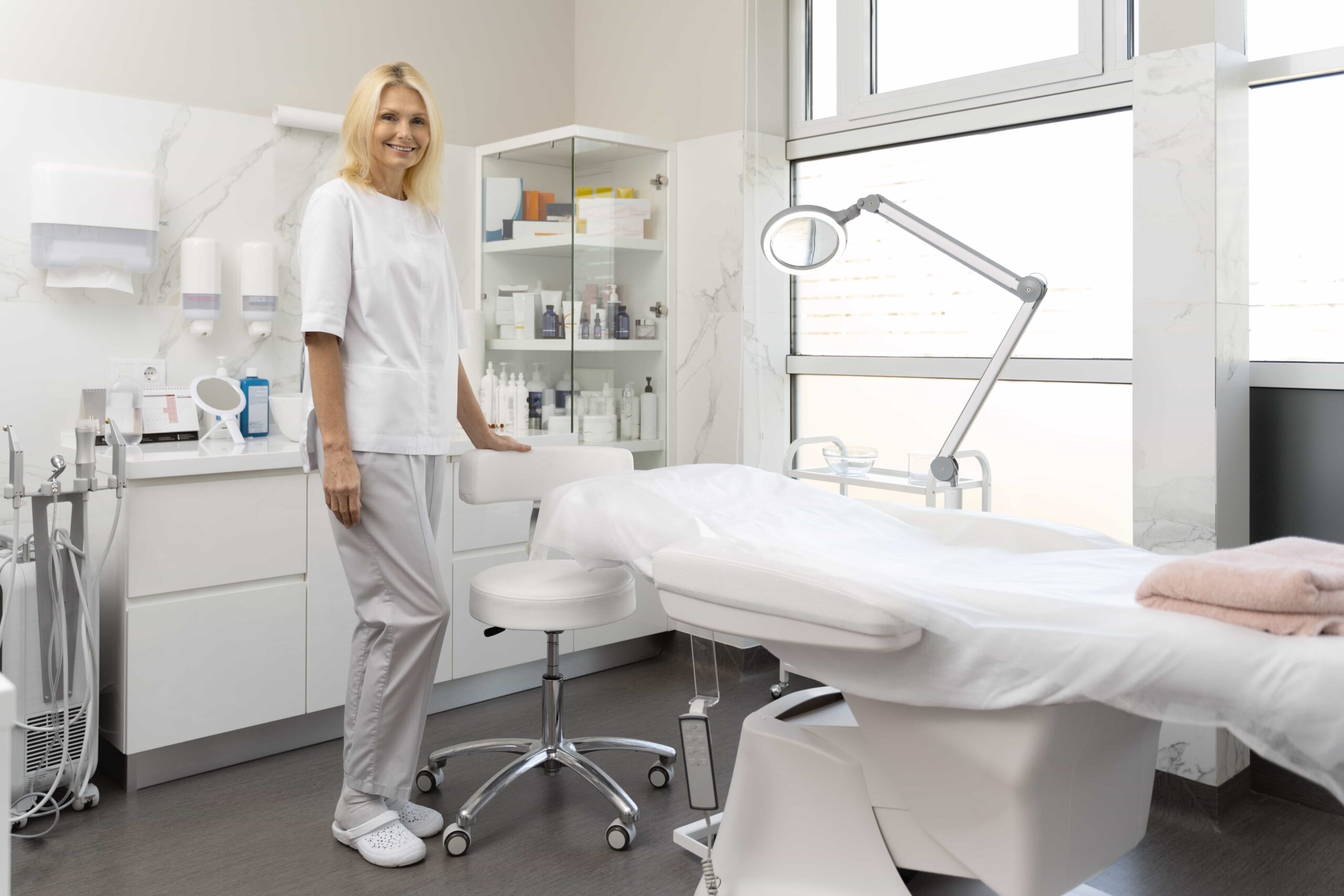 Smiling female aesthetician in a white uniform standing next to a clean treatment bed and magnifying lamp in a bright, modern cosmetic clinic room.