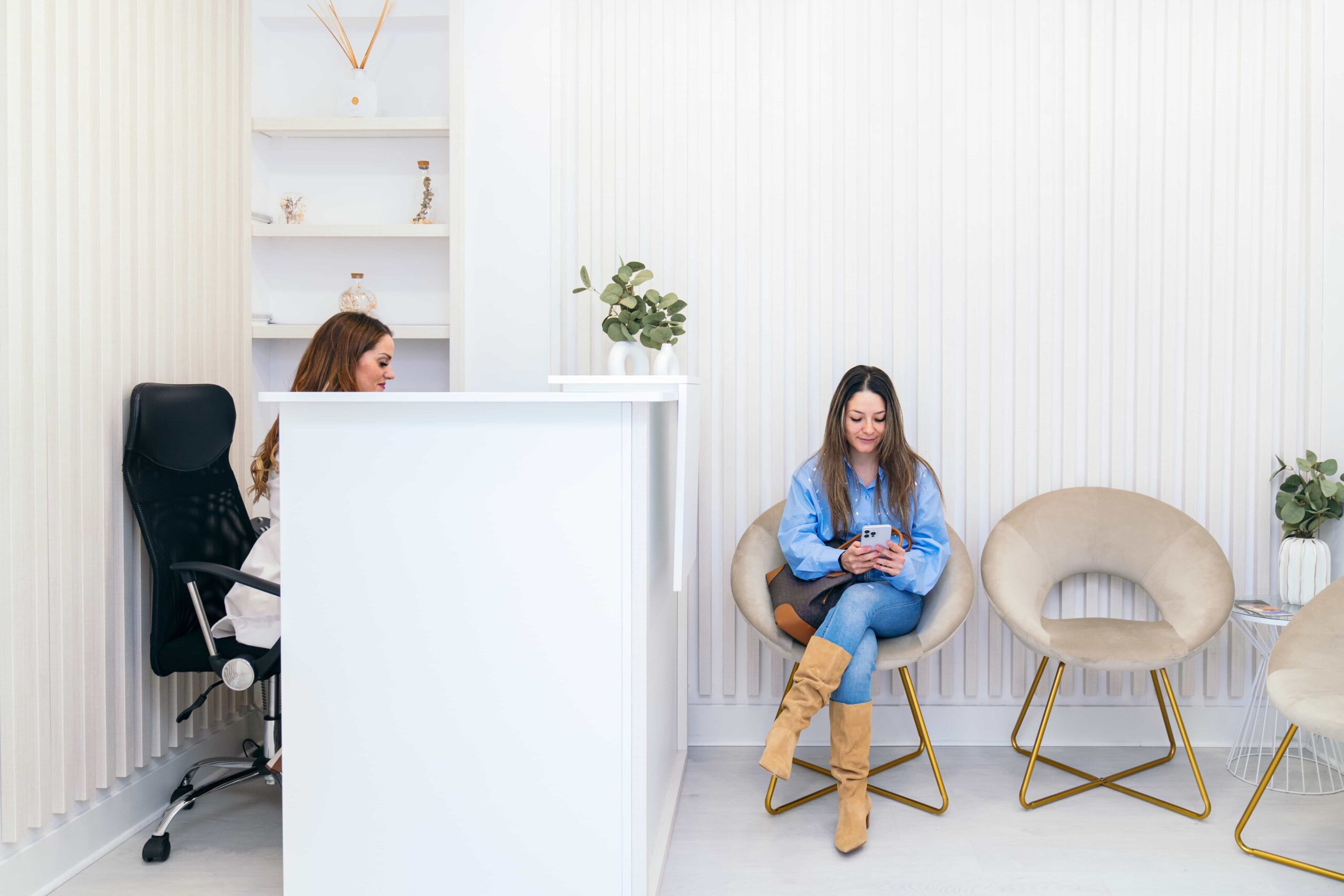 Bright and modern clinic waiting room with a white reception desk, featuring a patient sitting in a velvet chair checking her phone.