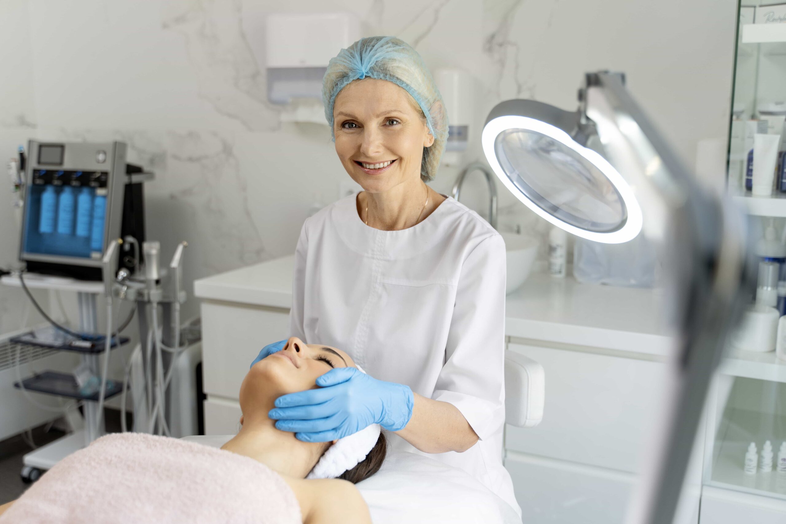 Smiling mature aesthetician wearing a hair net and blue gloves examining a client's face under a magnifying lamp in a professional treatment room.