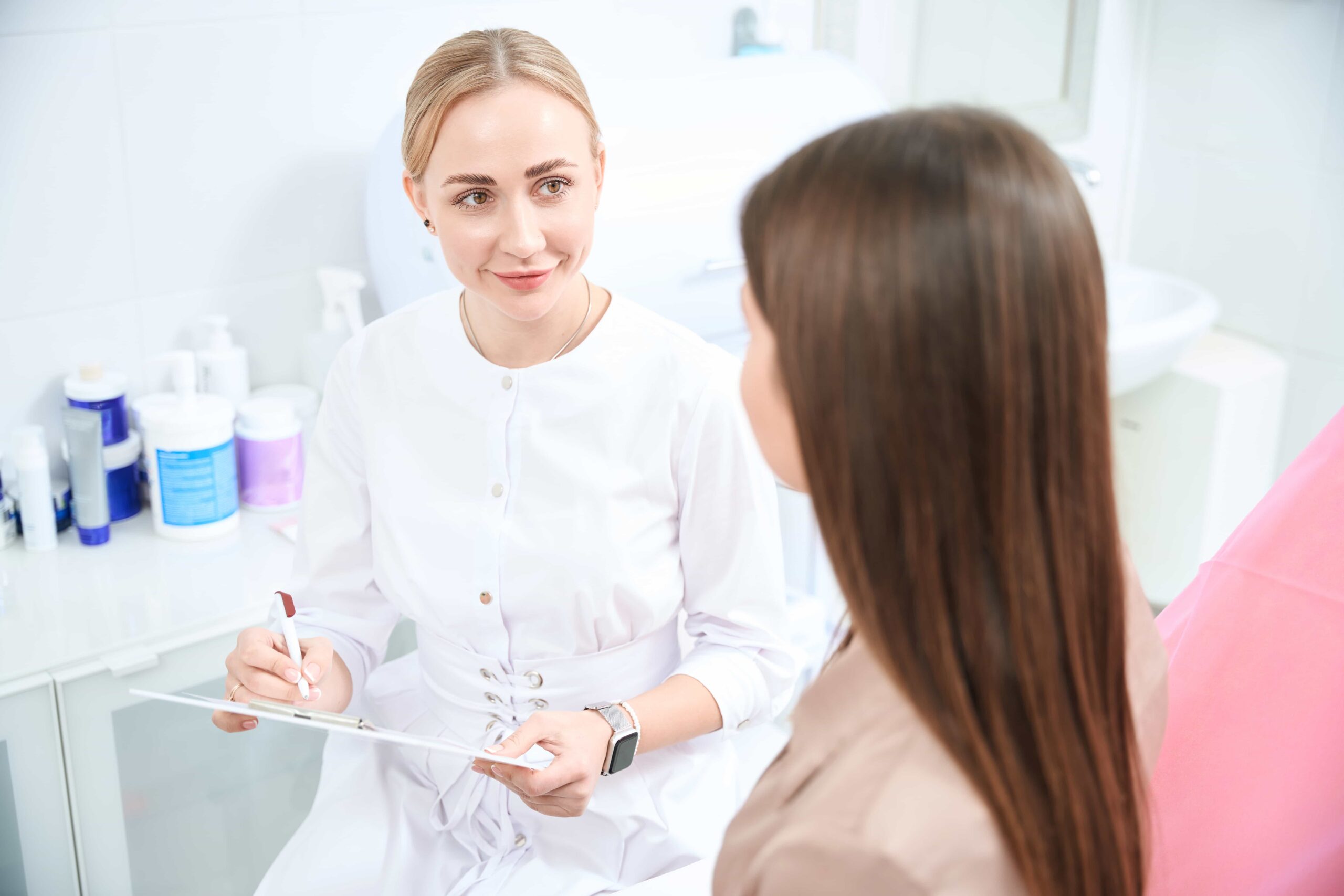Female aesthetician in a white uniform smiling while reviewing a form or consent document with a female client during a cosmetic consultation in a clinic.