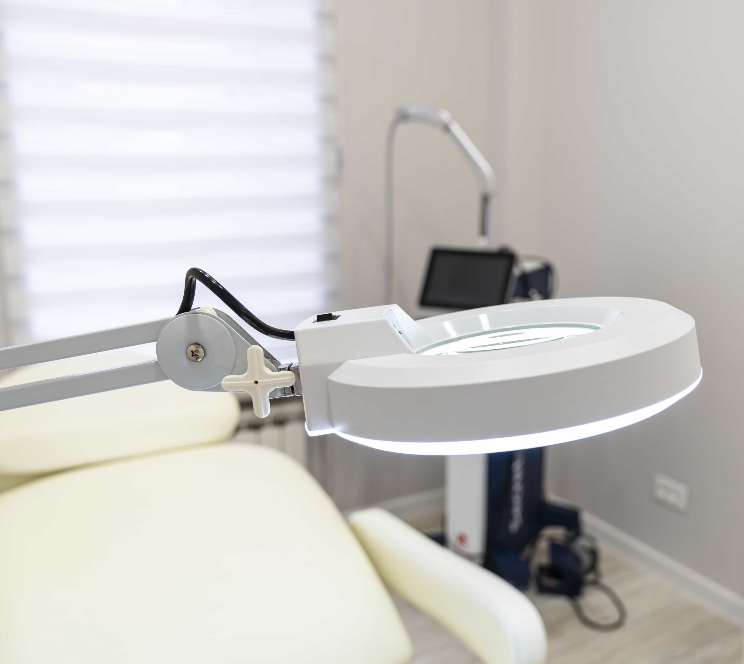 Close-up of a bright, circular magnifying lamp positioned over a clean treatment bed in a modern aesthetic clinic, with advanced cosmetic equipment visible in the background.