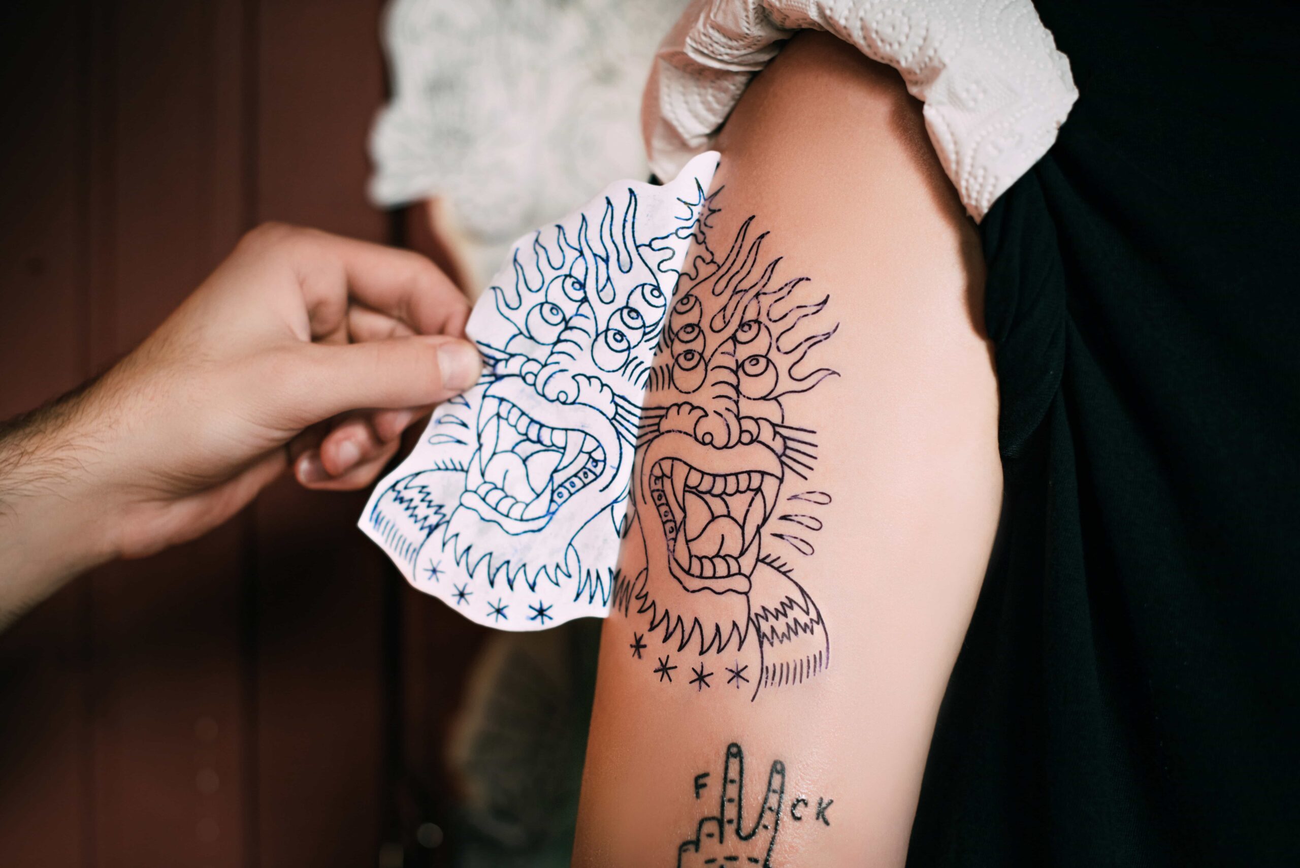 Close-up showing the temporary transfer outline of a traditional monstrous face tattoo applied to a person's upper arm next to the paper template, in preparation for inking.