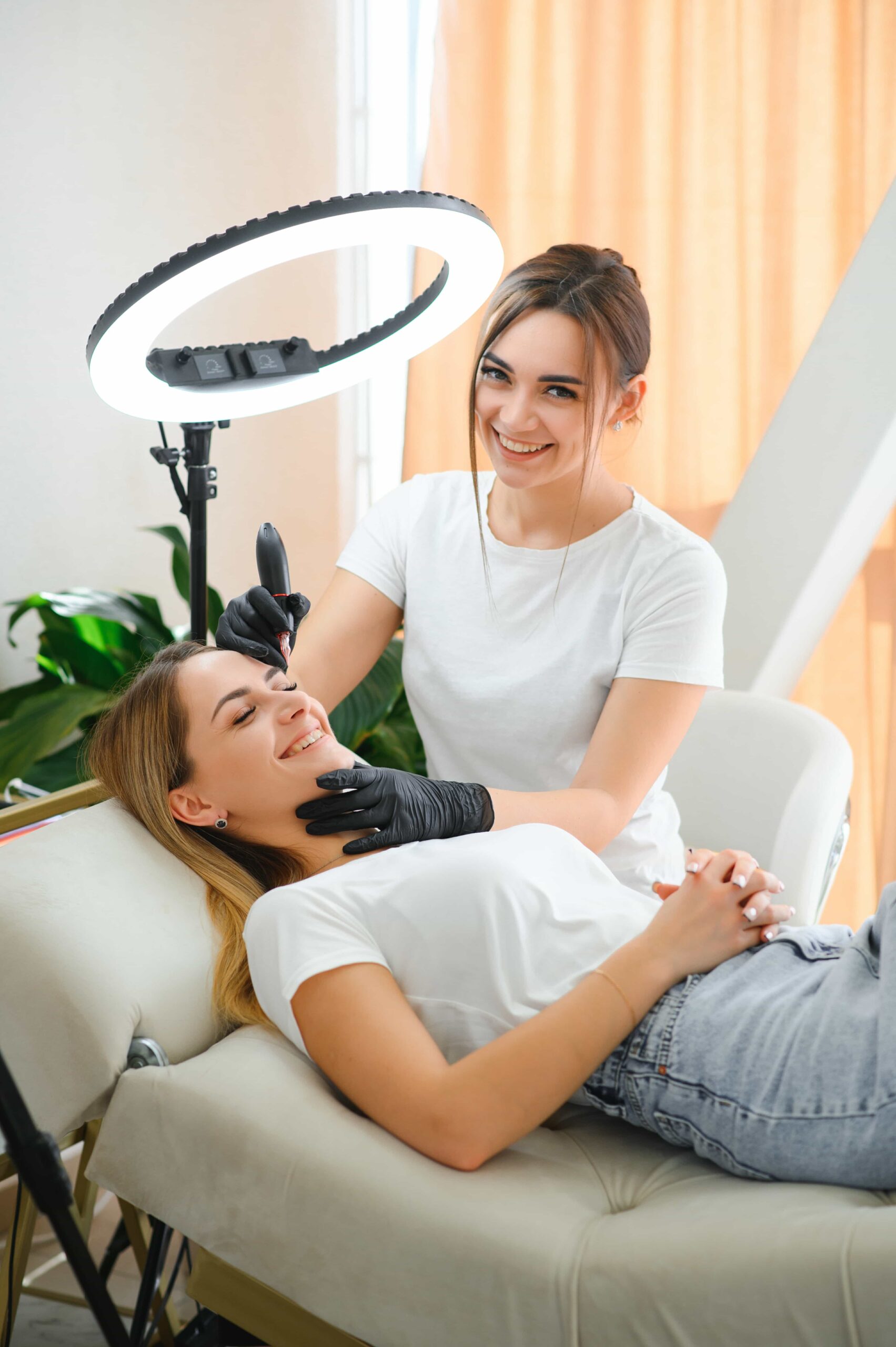 Smiling aesthetician in a white shirt and black gloves examining a client's jawline under a bright, circular ring light in a modern treatment room.