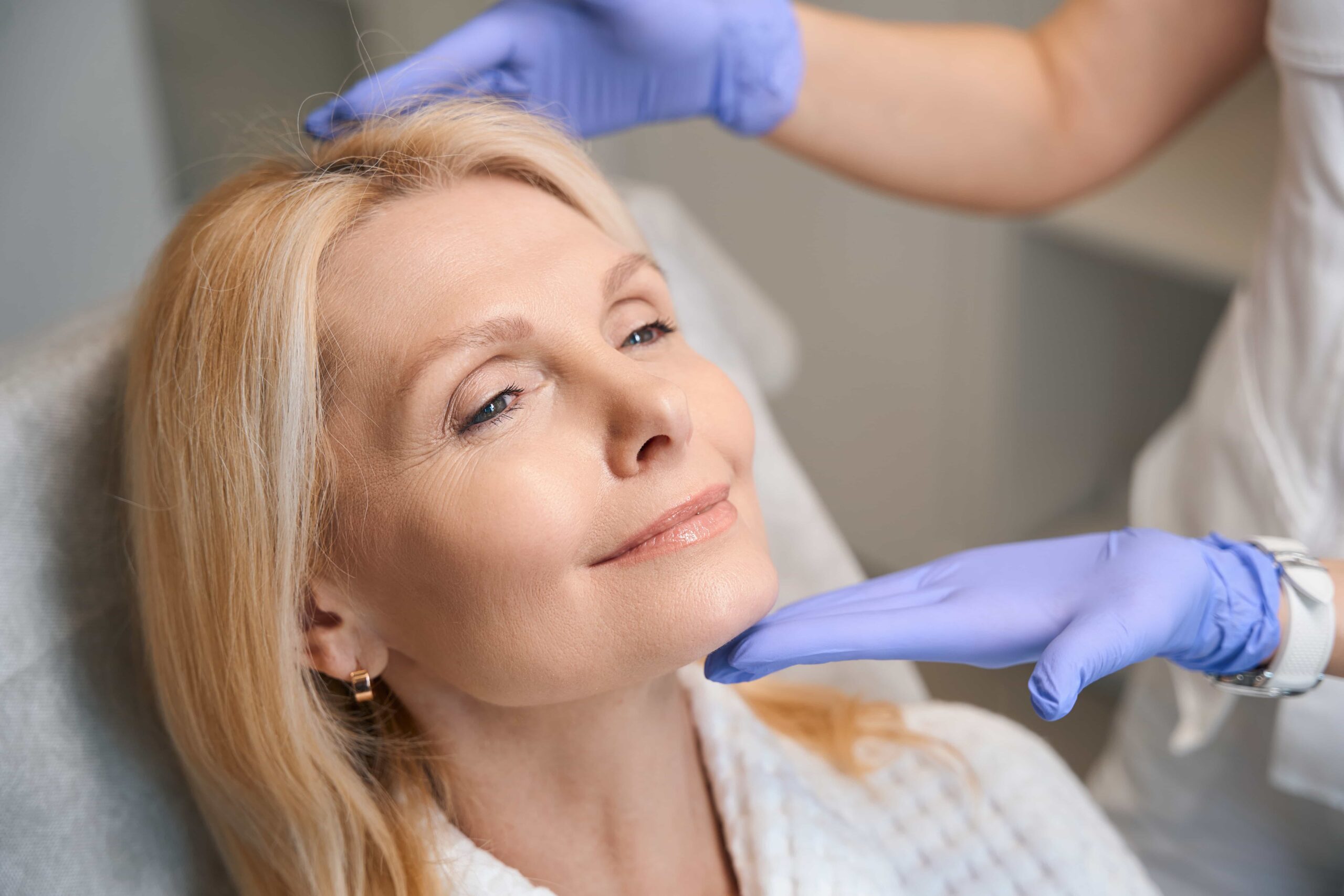 Smiling mature woman receiving a skin assessment or gentle facial massage, with a professional wearing blue gloves framing her chin and forehead.