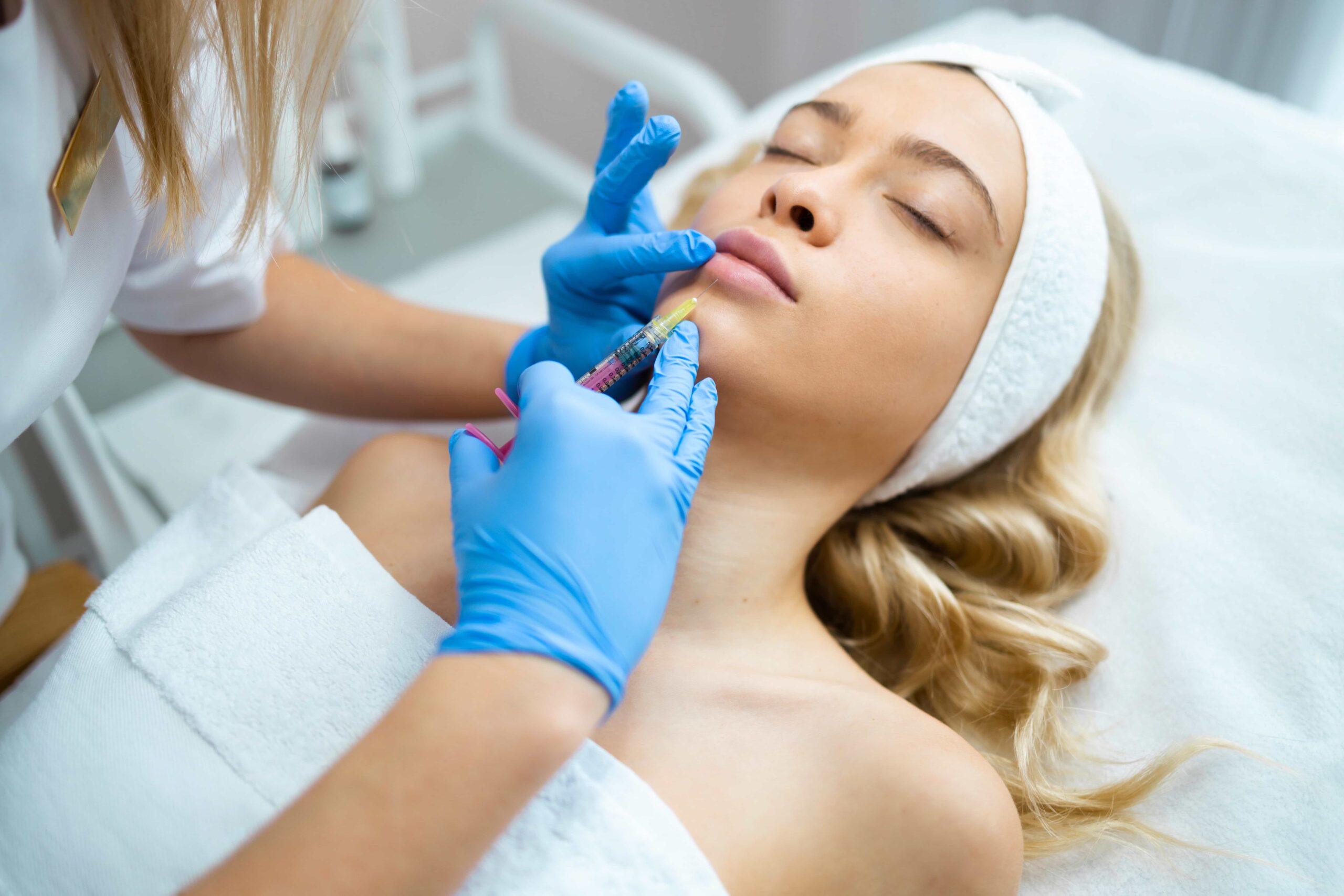 Young blonde woman receiving a cosmetic injection, likely lip filler, from a professional wearing blue gloves, while lying on a treatment bed.
