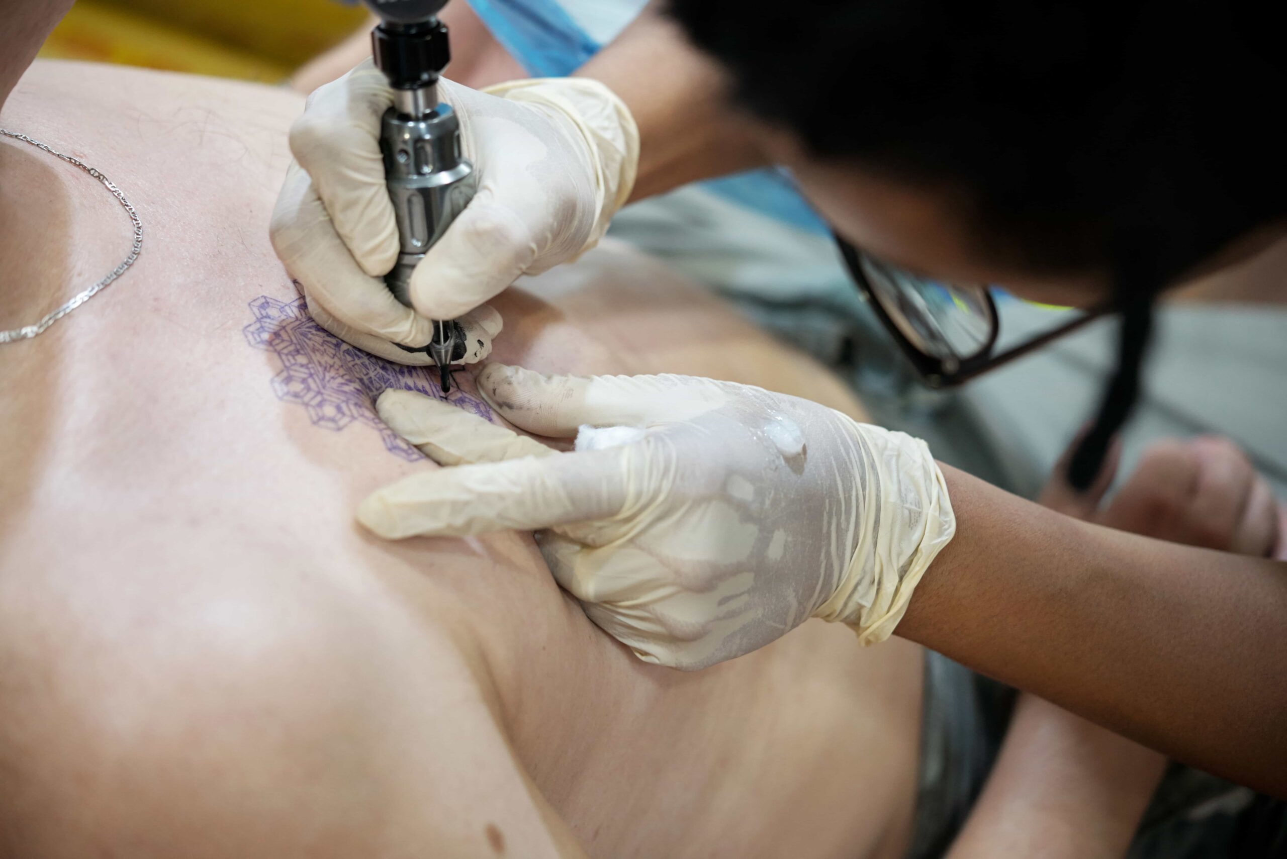 Close-up of a tattoo artist wearing white gloves using a tattoo machine to outline a purple design on a client's chest.