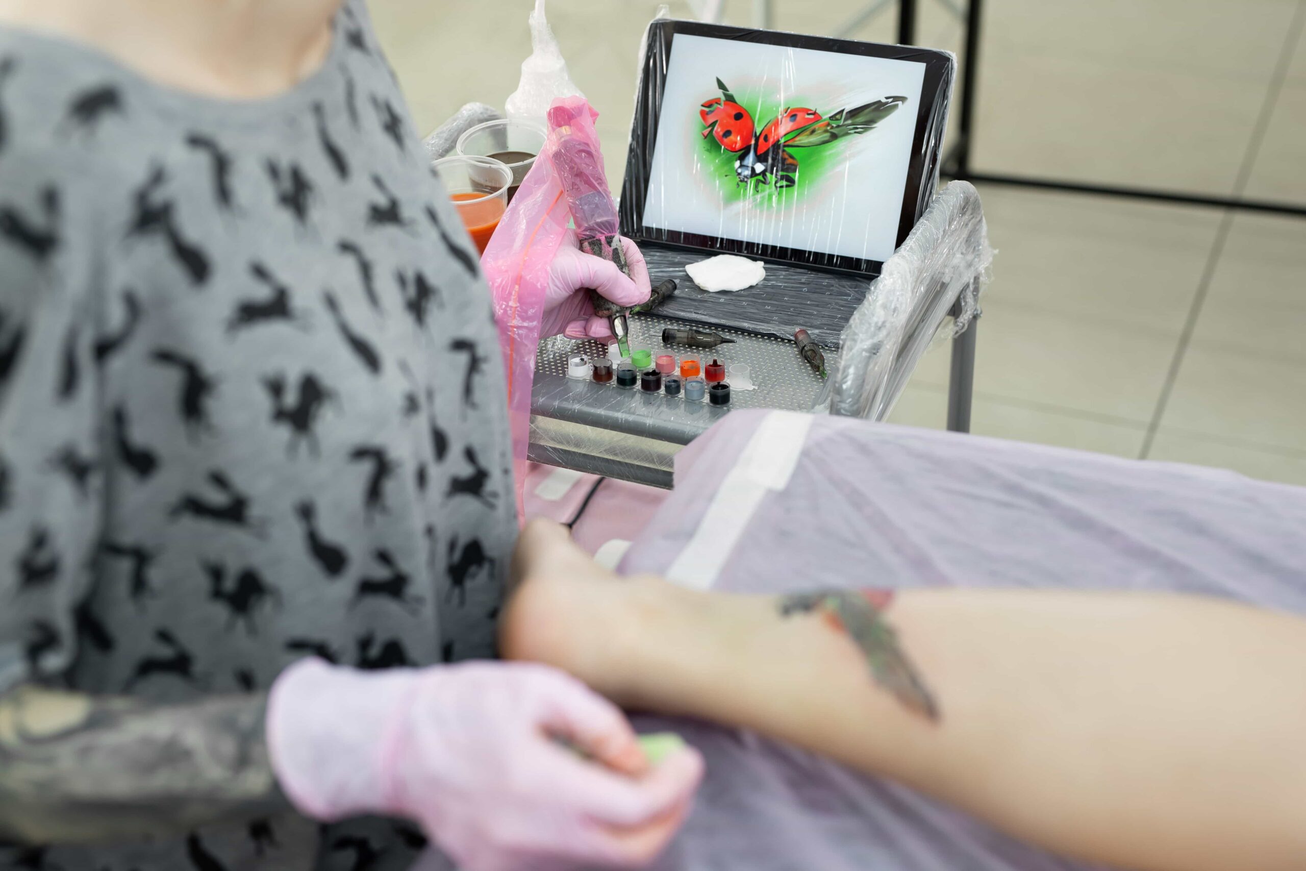 Tattoo artist wearing pink gloves working on a colorful tattoo on a client's leg, with the digital reference image of a ladybug visible on a tablet.