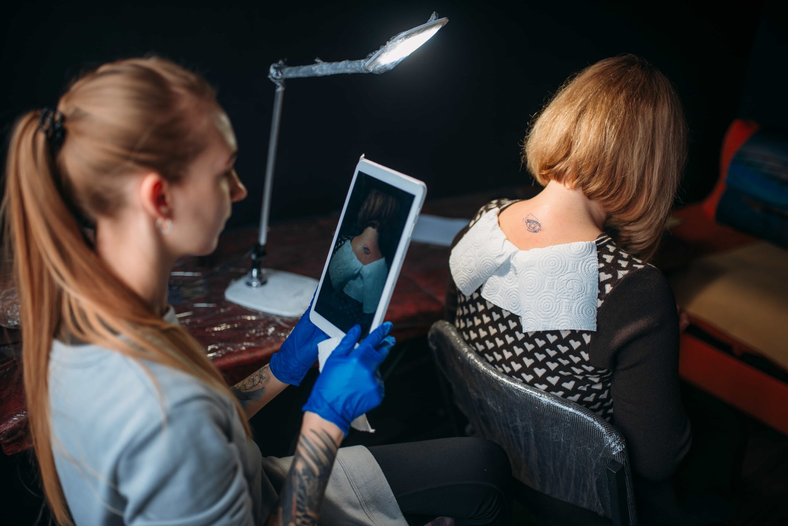 Female tattoo artist wearing blue gloves consulting a digital reference image on a tablet before working on a small black tattoo on the back of a client's neck.
