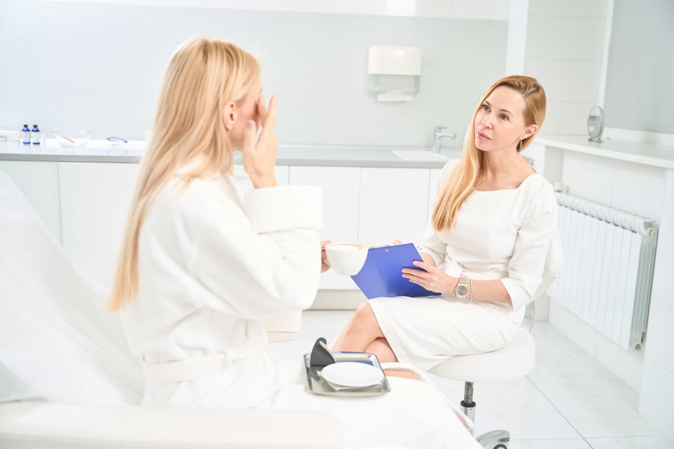 Female professional in a white dress listening intently to a blonde client who is touching her face while holding a cup, during a skincare consultation in a bright clinic.