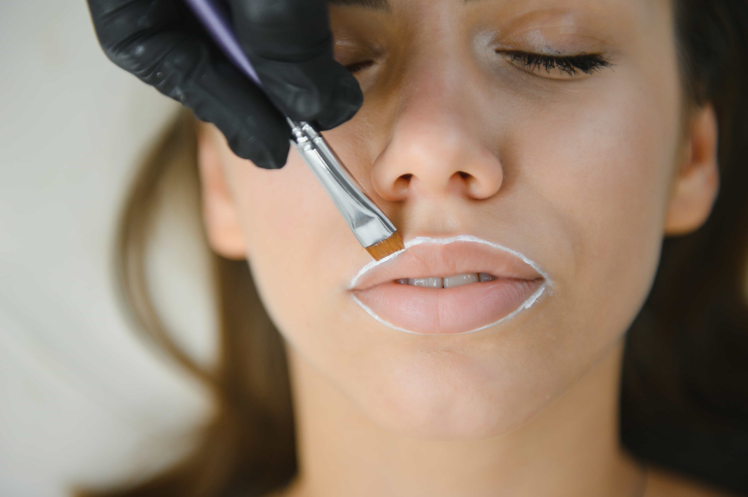 Close-up of a client receiving a treatment, with white outlining applied around her lips and an aesthetician in a black glove using a small brush near her mouth.