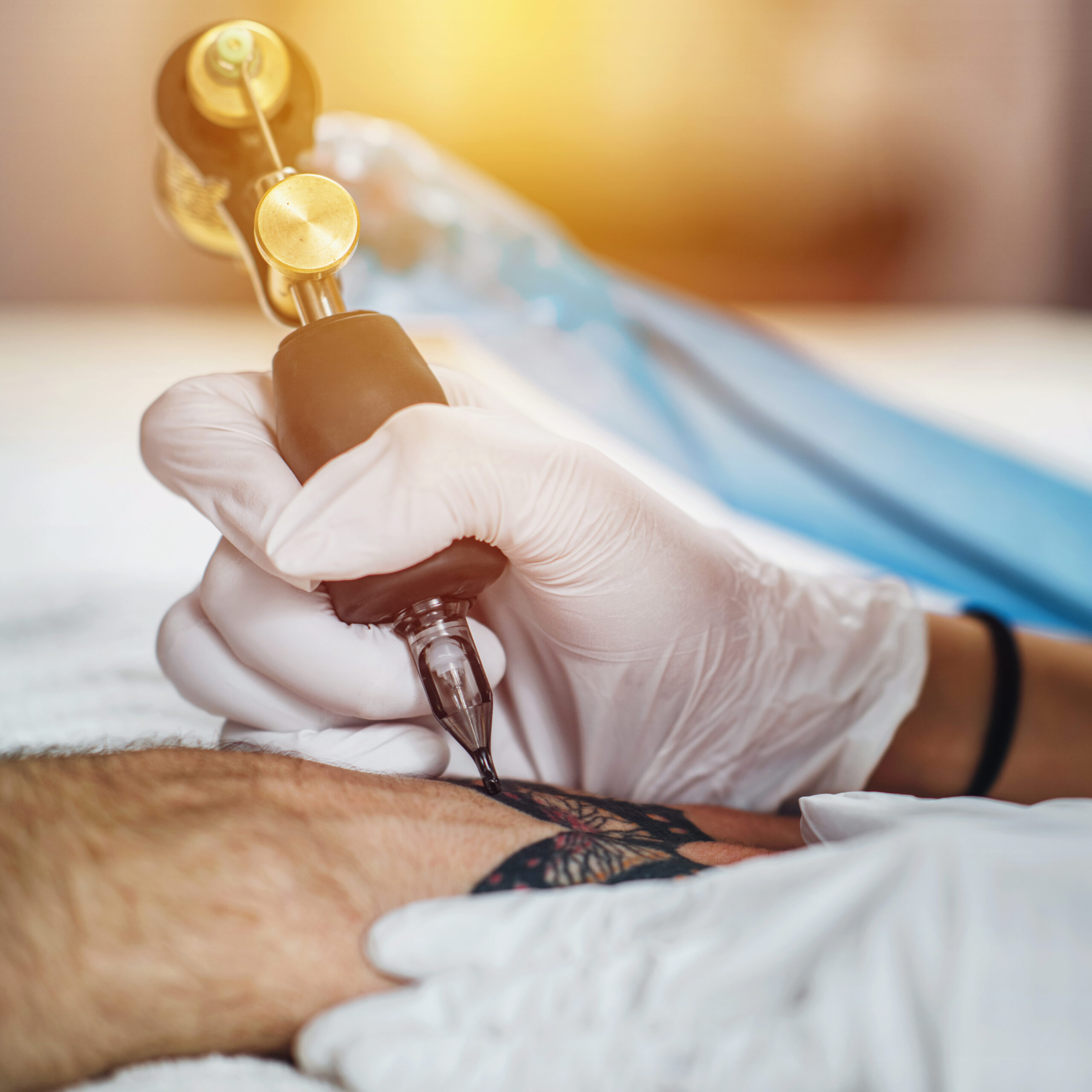 Close-up of a tattoo artist wearing white gloves using a tattoo machine to apply black ink to a tattoo design on a client's arm, with bright sunlight in the background.
