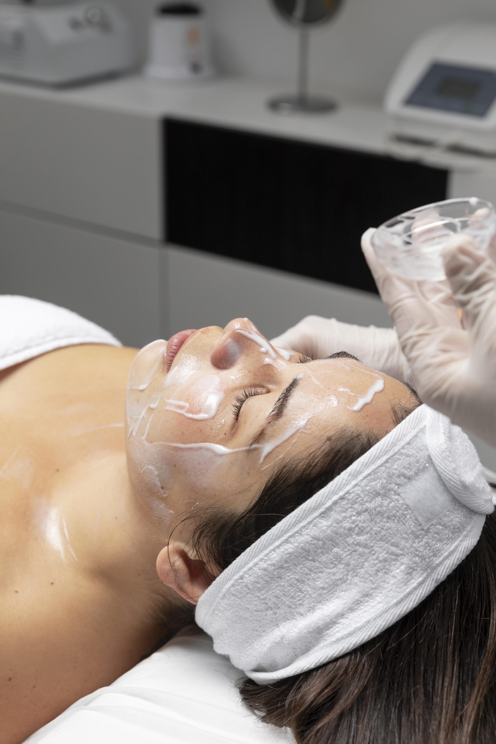 Close-up of an aesthetician wearing white gloves applying a thick, white cream or mask to a woman's face using a silicone brush during a facial.