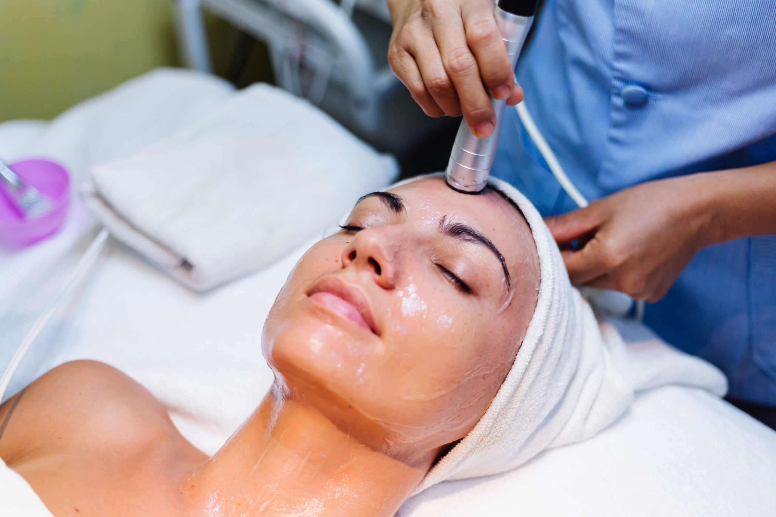 Woman receiving a non-invasive, device-based skincare treatment, such as ultrasound or radiofrequency, on her forehead by a professional aesthetician.