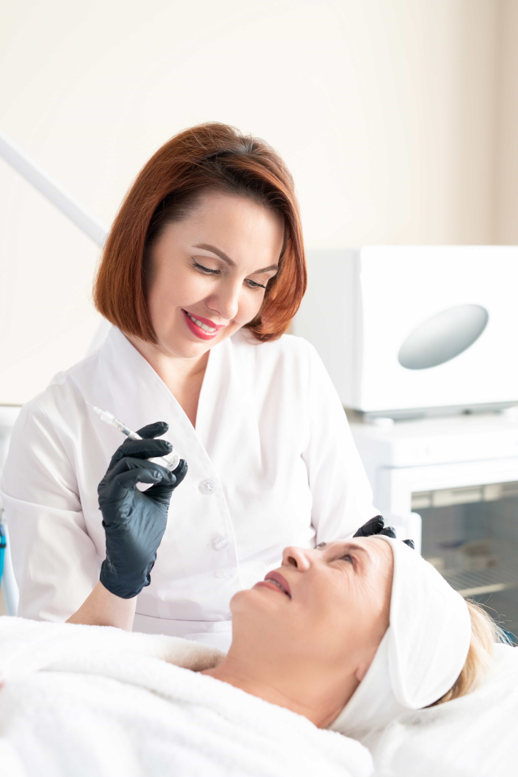 Smiling aesthetician holding an injection syringe, looking down at an older client who is prepared for a cosmetic treatment.