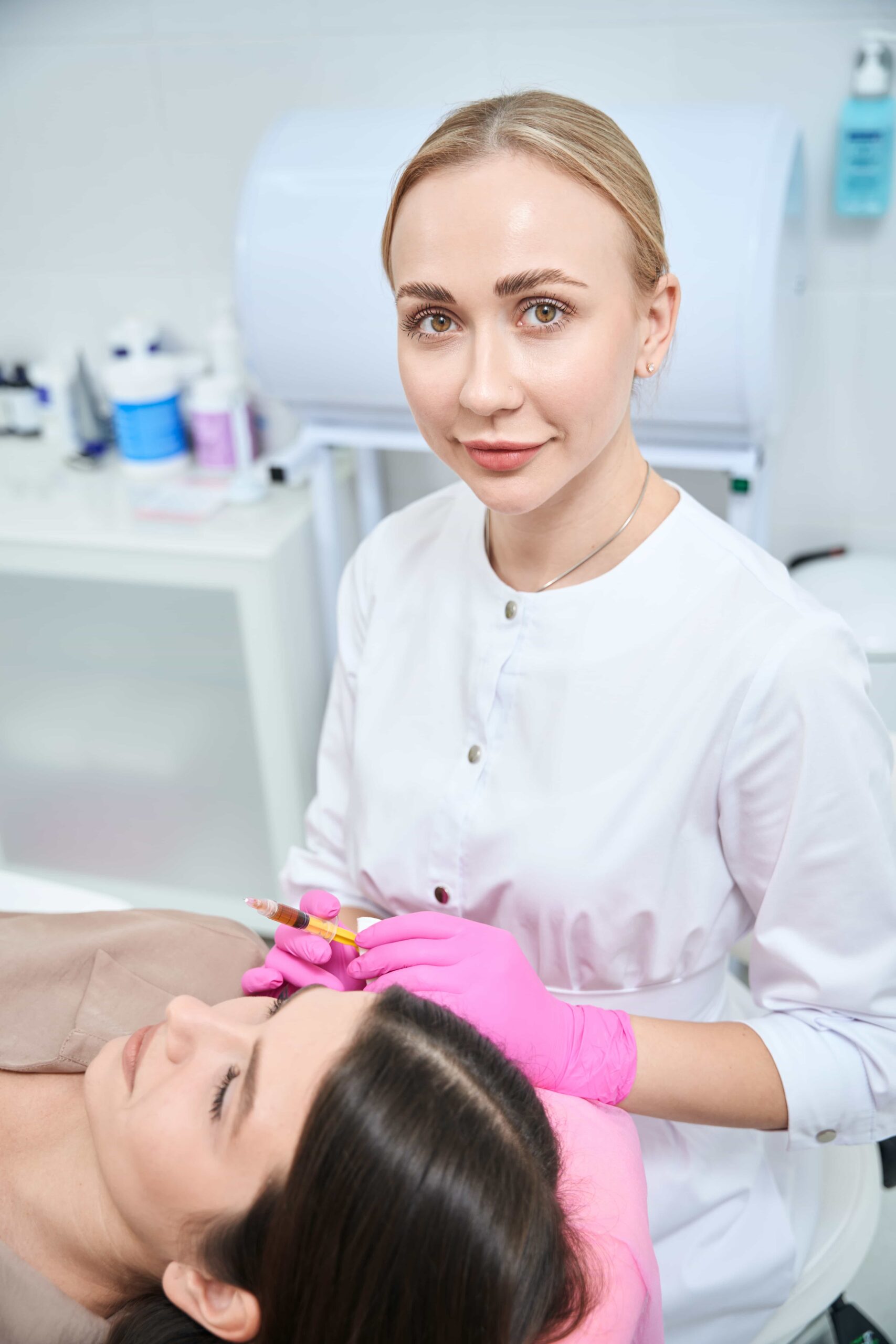 Female medical aesthetician in a white coat and pink gloves preparing to administer a facial injection, such as filler or Botox, to a client.
