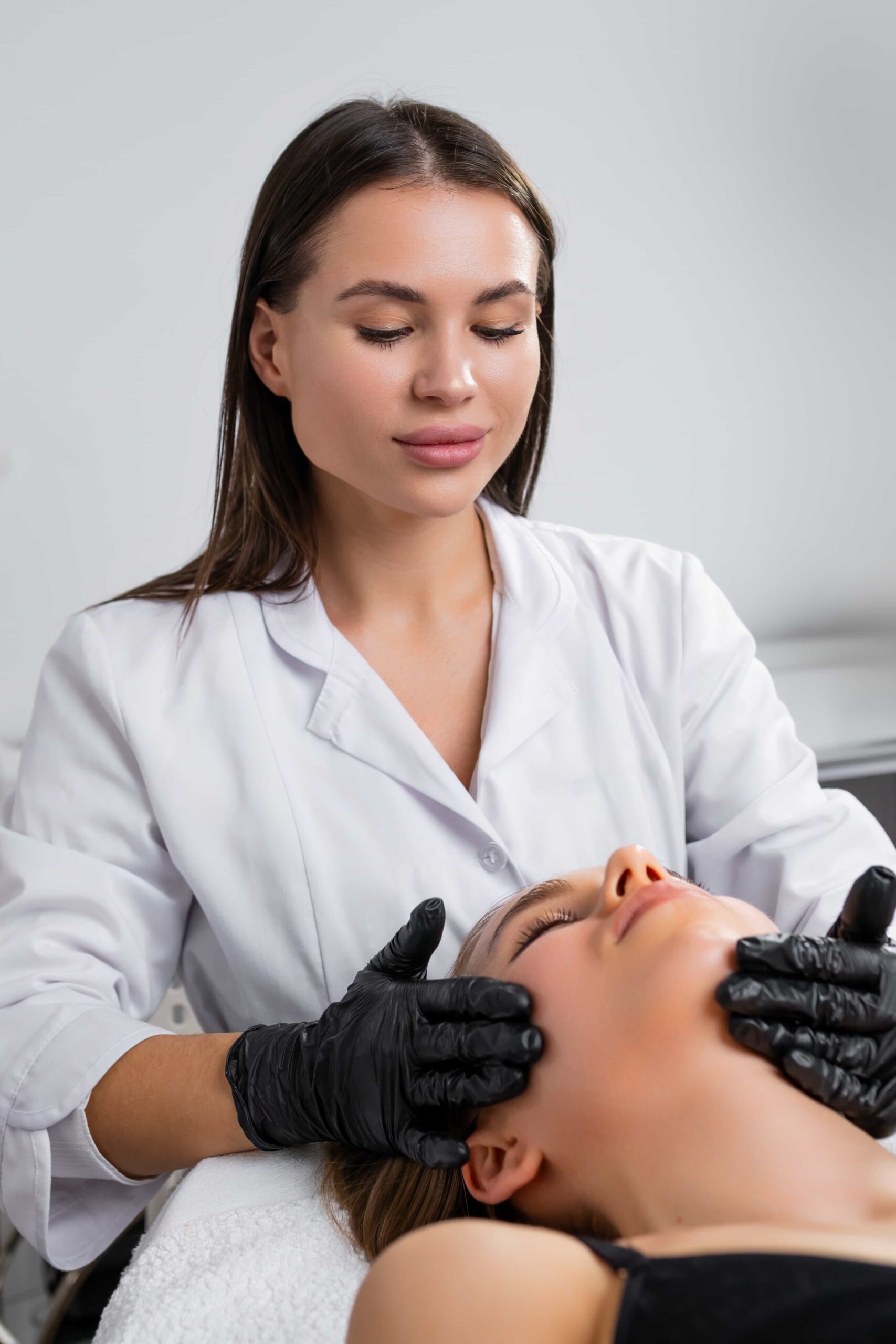 Professional aesthetician in a white lab coat and black gloves performing a soothing facial massage on a client's cheeks.